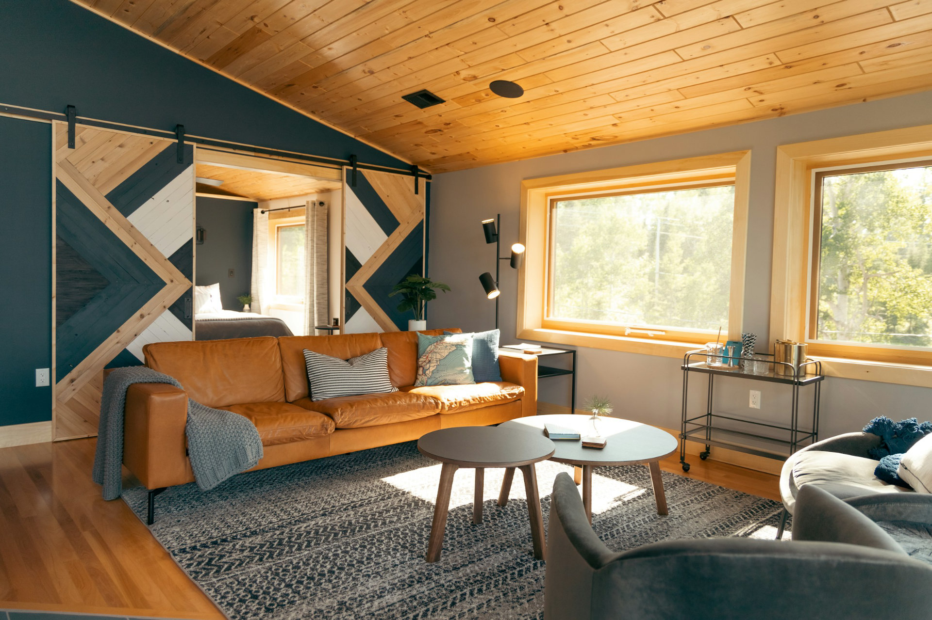 Photo de l'intérieur moderne et ensoleillé d'un chalet en montagne, avec des prédominantes de couleurs bleu foncé désaturé, bois et marron. Il y a un canapé, deux tables centrales et de belles grande fenêtres